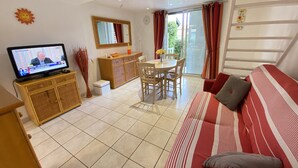 A living room with a television, dining table, chairs, a sofa, and a balcony with curtains. - Air-conditioned pavilion with pool, near beach, 2 bedrooms (Valras-Plage)
