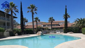 A swimming pool with a stone deck, surrounded by palm trees and a building in the background. - Air-conditioned pavilion with pool, near beach, 2 bedrooms (Valras-Plage)