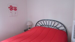 A bedroom with a metal headboard, a red bedspread, a nightstand with a lamp, and a wall-mounted picture of flowers. - Air-conditioned pavilion with pool, near beach, 2 bedrooms (Valras-Plage)