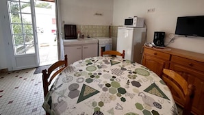 A compact kitchen with a refrigerator, microwave, and sink. There's a dining table with a geometric-patterned tablecloth and wooden chairs. A television is mounted on the wall.