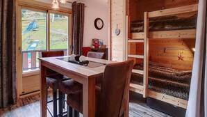 A cozy room with a wooden dining table, chairs, a clock, and a window with a view of a green landscape. - La Clusaz: Studio 4 pers, Balcony, Wifi, Pets allowed (La Clusaz)