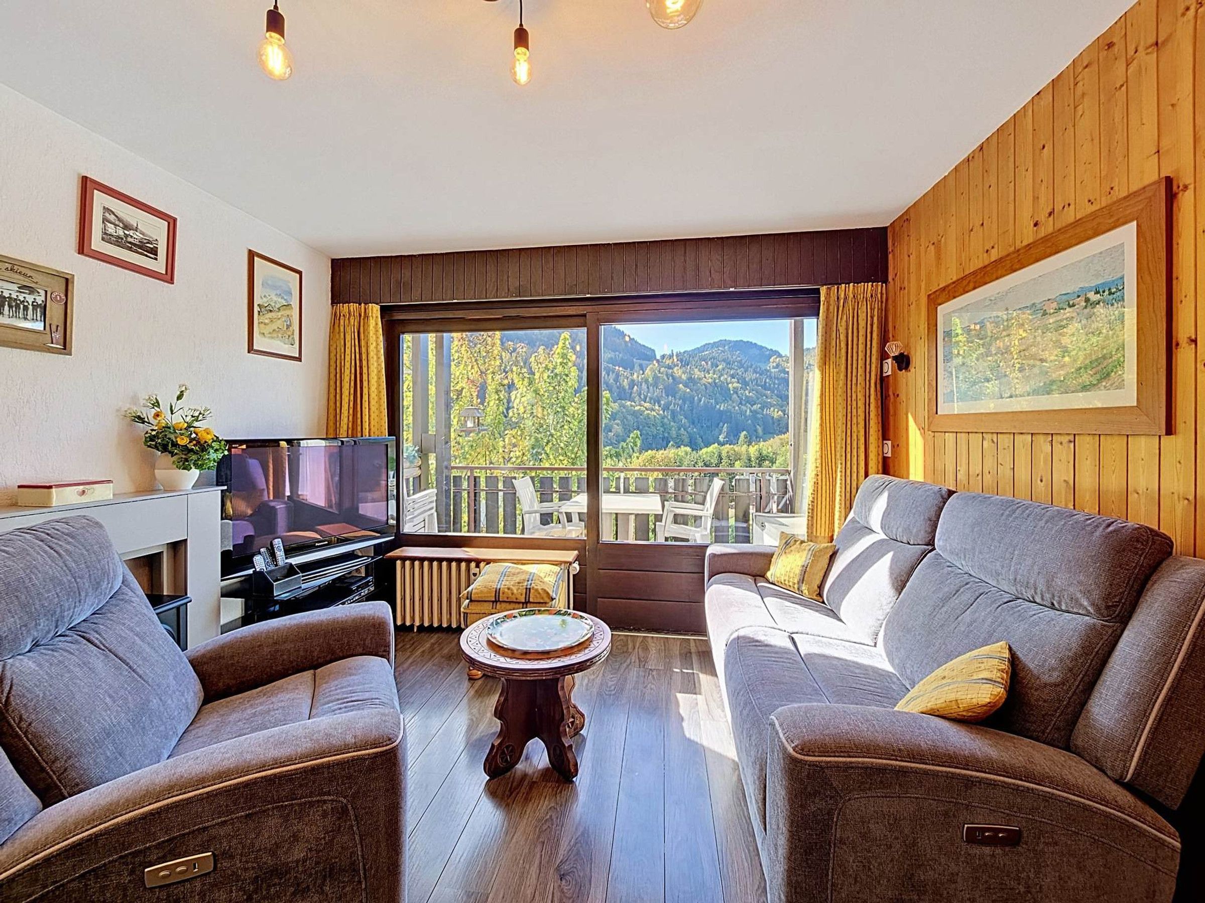 A living room with a television, two sofas, a coffee table, and a view of a mountainous landscape through large windows.