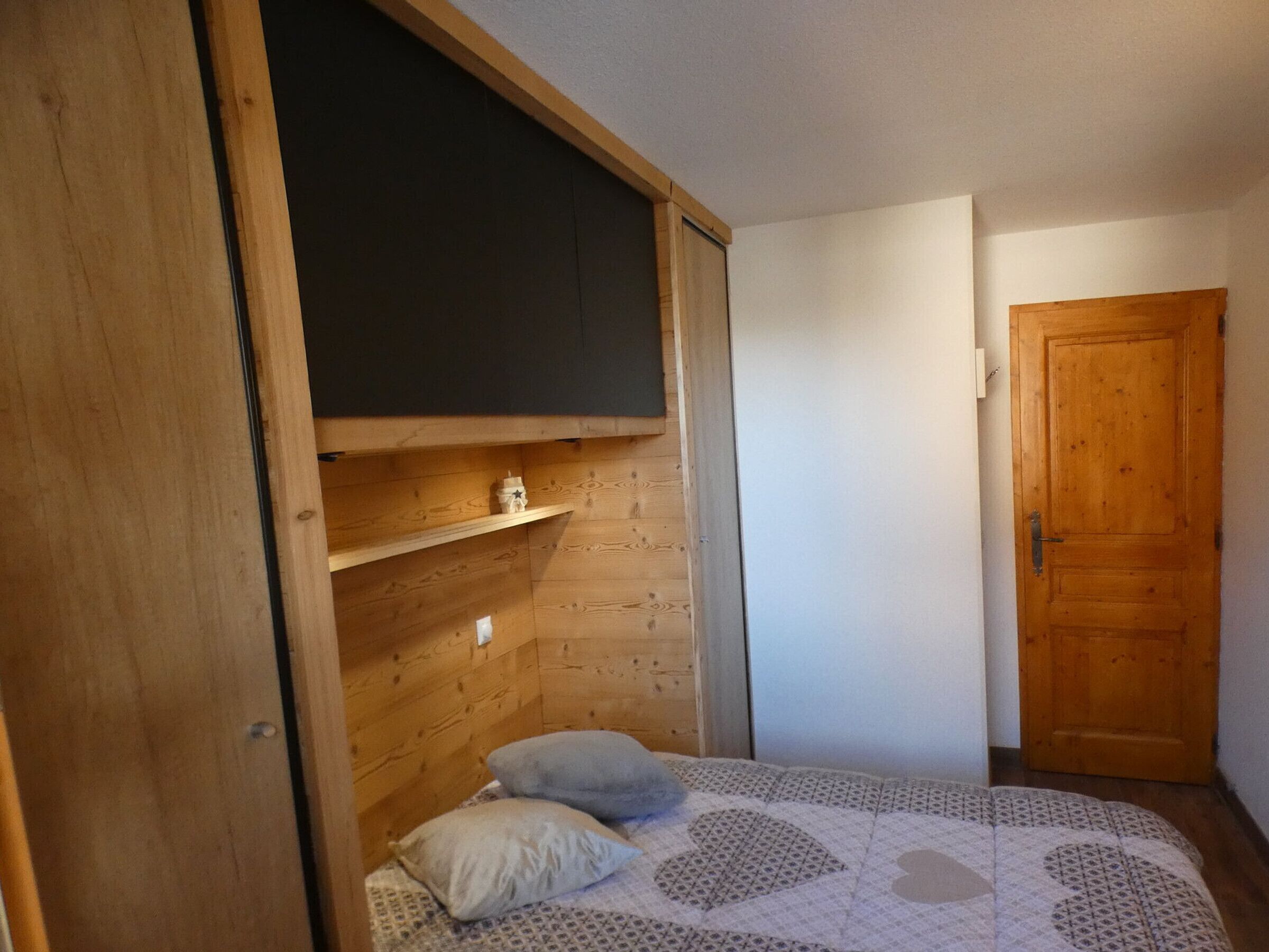 Une petite chambre à coucher accueillante, avec un plafond en pente en bois, un lit simple recouvert d’une courtepointe à motifs, deux oreillers et une petite étagère sur laquelle est posé un objet décoratif.