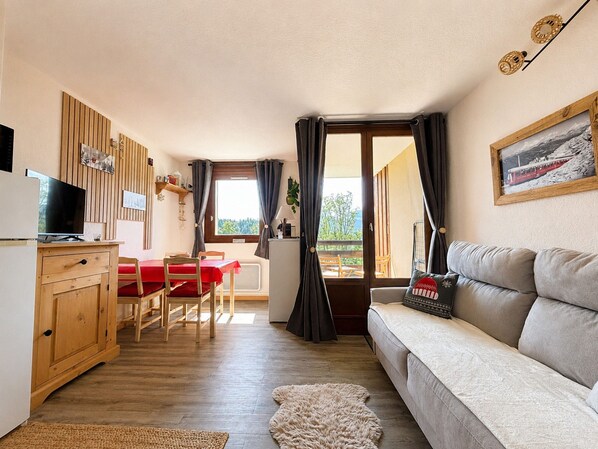 A cozy living room with a sofa, a TV, a dining table with chairs, and a kitchen area with a fridge.