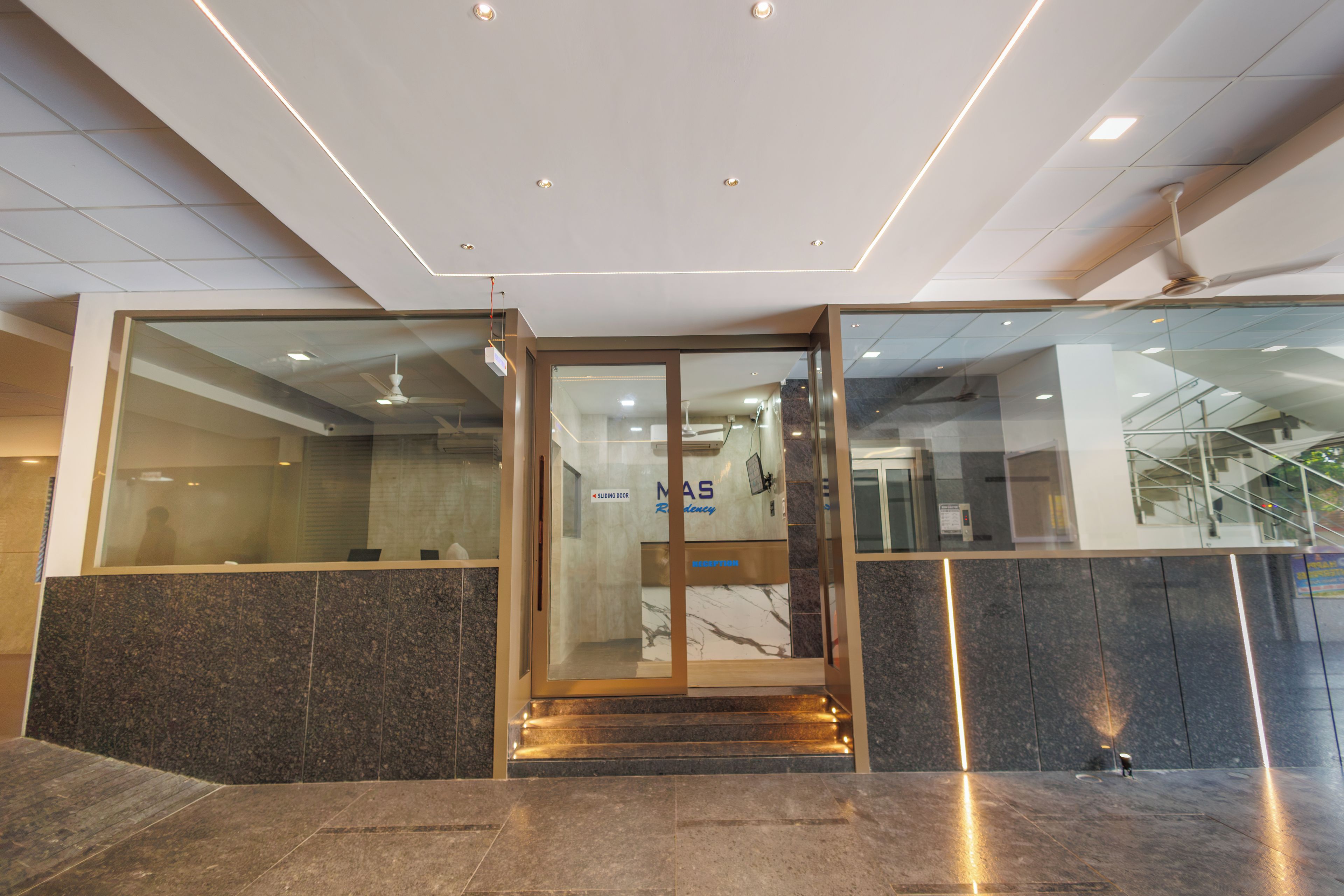 Zona de recepción de un hotel con entrada por puerta de cristal, un mostrador de recepción y una escalera que sube a un piso superior.