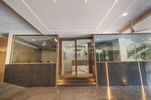 Zona de recepción de un hotel con entrada por puerta de cristal, un mostrador de recepción y una escalera que sube a un piso superior.