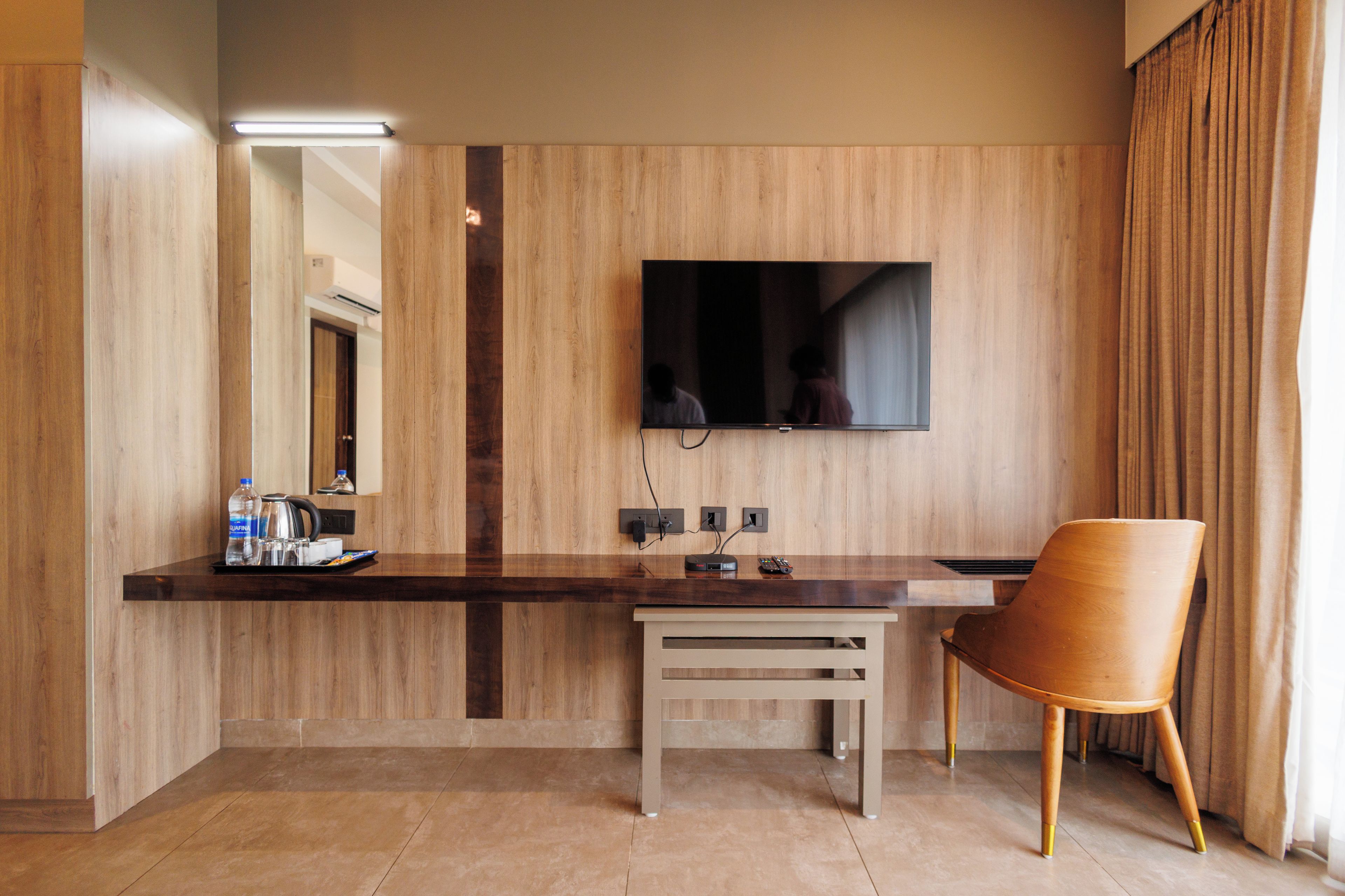 A hotel room with a wooden panel wall, a flat-screen TV mounted above a wooden desk, a chair, and a small table.