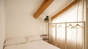 Un dormitorio con cama, lámpara colgante, vigas de madera, ventana con cortinas y un perchero.