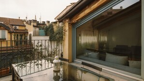 Un balcón con mesa y sillas de vidrio, vistas a la ciudad y un ventanal con un sofá en el interior.