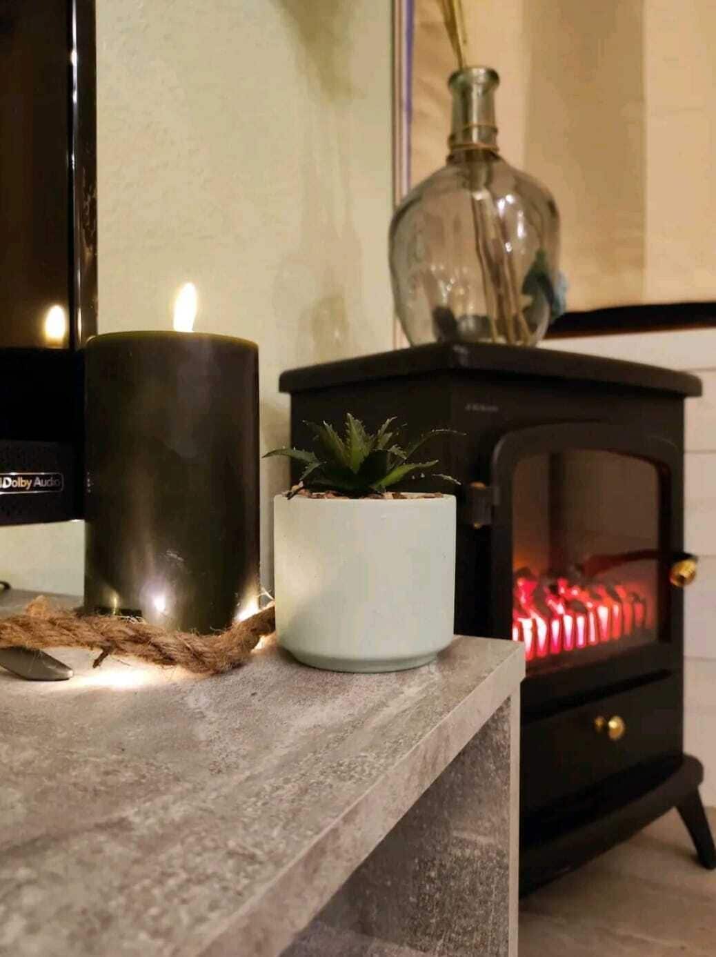 Eine gemütliche Ecke mit brennender Kerze, einer kleinen Topfpflanze, einem Kamin und einem Fernseher.