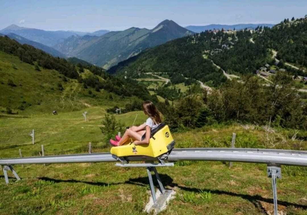 Une personne est assise sur une sorte de luge jaune et descend une pente herbeuse, avec une montagne en arrière-plan.