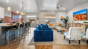 A modern living room with a blue sofa, a dining area with bar stools, and a kitchen with a white countertop. - Zion Village 251|Private Pool, Xbox, Grill, Resort Amenities, and more! (Hurricane)