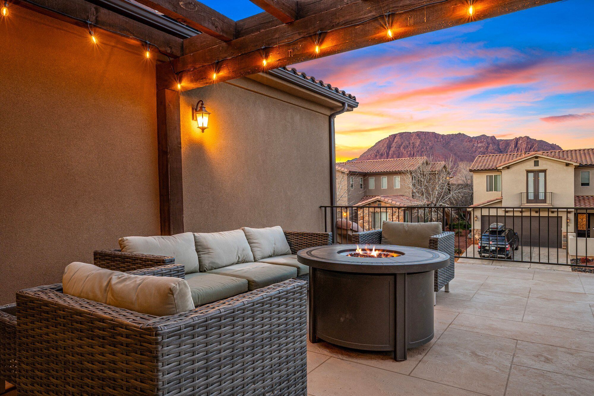 A patio with wicker furniture, a round table with a fire pit, string lights, and a view of mountains at sunset.