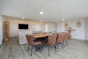 A modern dining area with a wooden table, brown chairs, a white sofa, and a mounted flat-screen TV. - Volcanoa: Holiday Home with Ocean & Volcano Views (Villaverde)