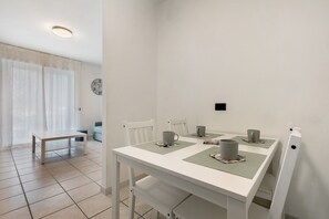 Un coin repas avec une table blanche, quatre chaises et un banc. Une horloge est accrochée au mur, et une fenêtre avec des rideaux est visible.