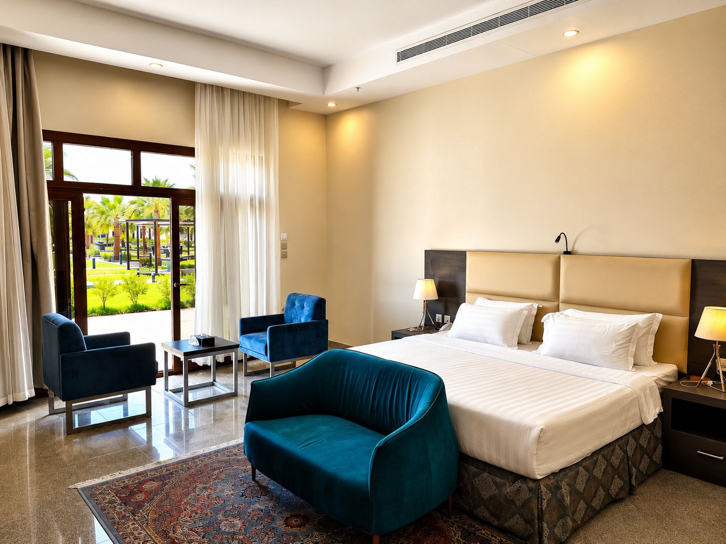 A hotel room with a large bed, a desk, two chairs, a nightstand with a lamp, and a view of a garden through the window.