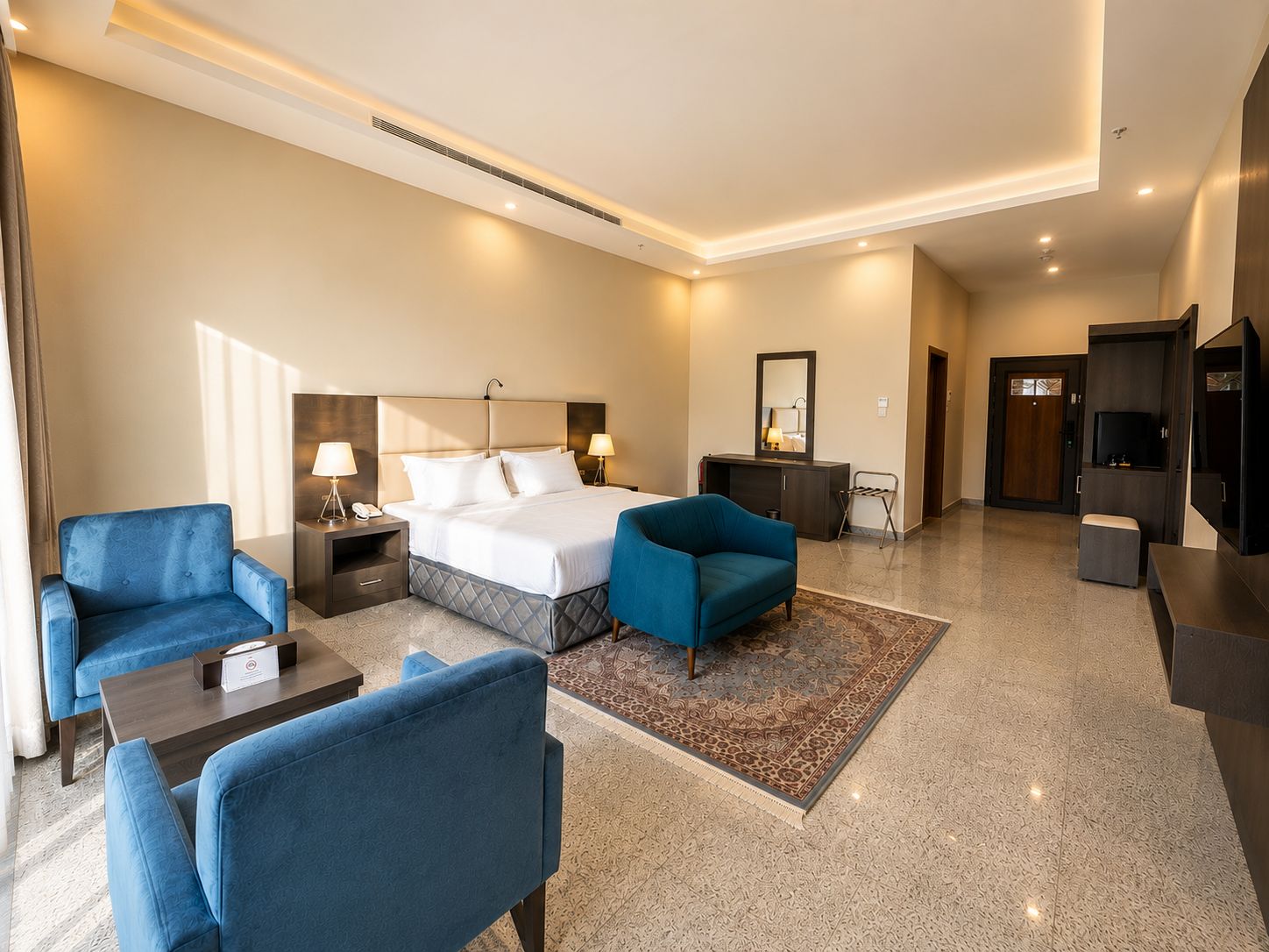 A hotel room with a bed, two armchairs, a small table, a desk, a TV, and a mirror.