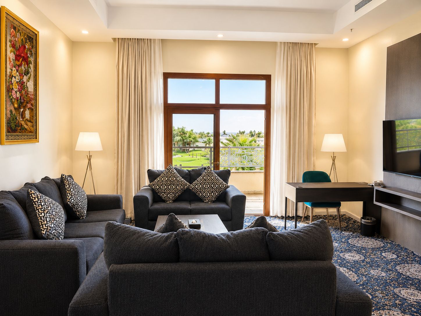 A hotel room with a sofa set, a desk, a television, and a view of a landscaped area.