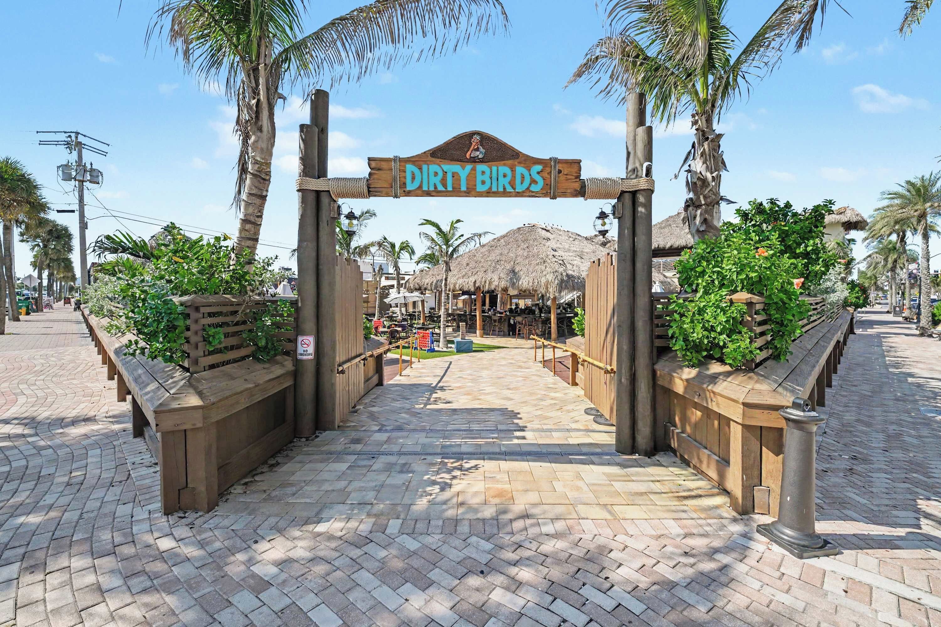 A wooden entrance gate with a sign reading 'DIRTY BIRDS' leading to a thatched-roof structure.
