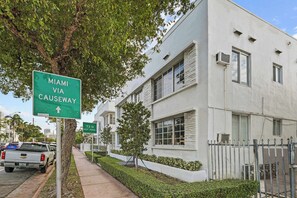 Un edificio residencial con fachada blanca, una calle bordeada de árboles y una señal verde direccional que indica 'Miami Via Causeway'.