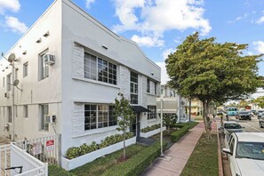 A modern white building with large windows, a gated driveway, and a tree-lined sidewalk.
