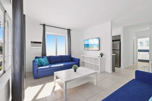 Una sala de estar moderna con un sofá azul, una mesa de centro blanca y vista al mar a través de amplios ventanales.