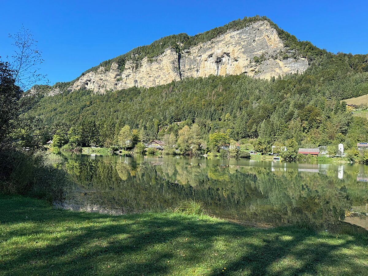 Un lago tranquilo con montañas al fondo, rodeado de abundante vegetación y algunos edificios.