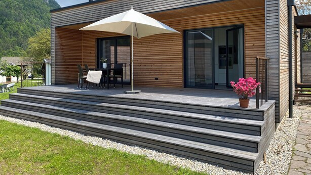 Een modern houten huis met een overdekte patio, buitenmeubilair en uitzicht op een bergachtig landschap.