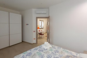 A bedroom with a bed, a wardrobe, an air conditioning unit, and a dining area in the background.
