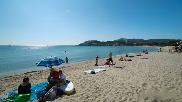 A beach with people sunbathing, a blue umbrella, and a sailboat in the distance.