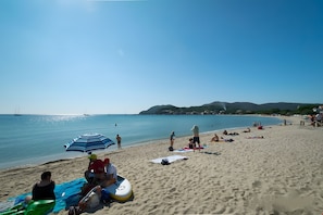 A beach with people sunbathing, a blue umbrella, and a sailboat in the distance.