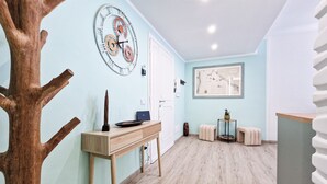 A hallway with a wooden tree-like structure, a wall clock, a framed map, a desk with a laptop, and a small round table with a plant.