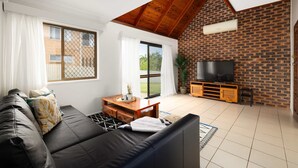 A living room with a black leather sofa, a wooden coffee table, a TV on a brick wall, and a sliding glass door leading to an outdoor area.
