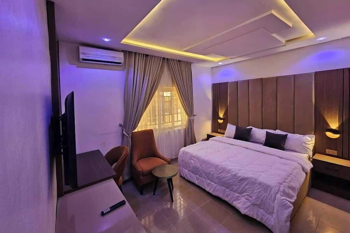 A modern hotel room with a large bed, a desk, and a chair. There is a TV on the wall and a window with curtains.