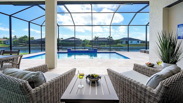 Un espace aménagé au bord de la piscine, avec des meubles en osier, une table sur laquelle se trouve un saladier rempli de fruits et un verre de vin, et une vue sur la piscine et sur des maisons situées au loin.