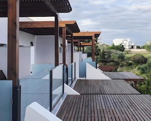 A modern outdoor deck with wooden flooring and glass railings, overlooking a landscaped area with distant buildings.