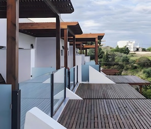 A modern outdoor deck with wooden flooring and glass railings, overlooking a landscaped area with distant buildings.