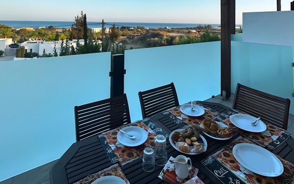 A table set for breakfast with plates, cups, and pastries, overlooking a coastal landscape.