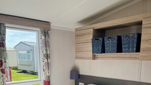 A neatly made bed with white linens, a bedside lamp, and a view of a grassy area outside. - Modern caravan, 5 mins walk to sandy beaches and South West coast path (Padstow)