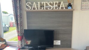 A living room with a television, a fireplace, and a shelf with board games. - Modern caravan, 5 mins walk to sandy beaches and South West coast path (Padstow)