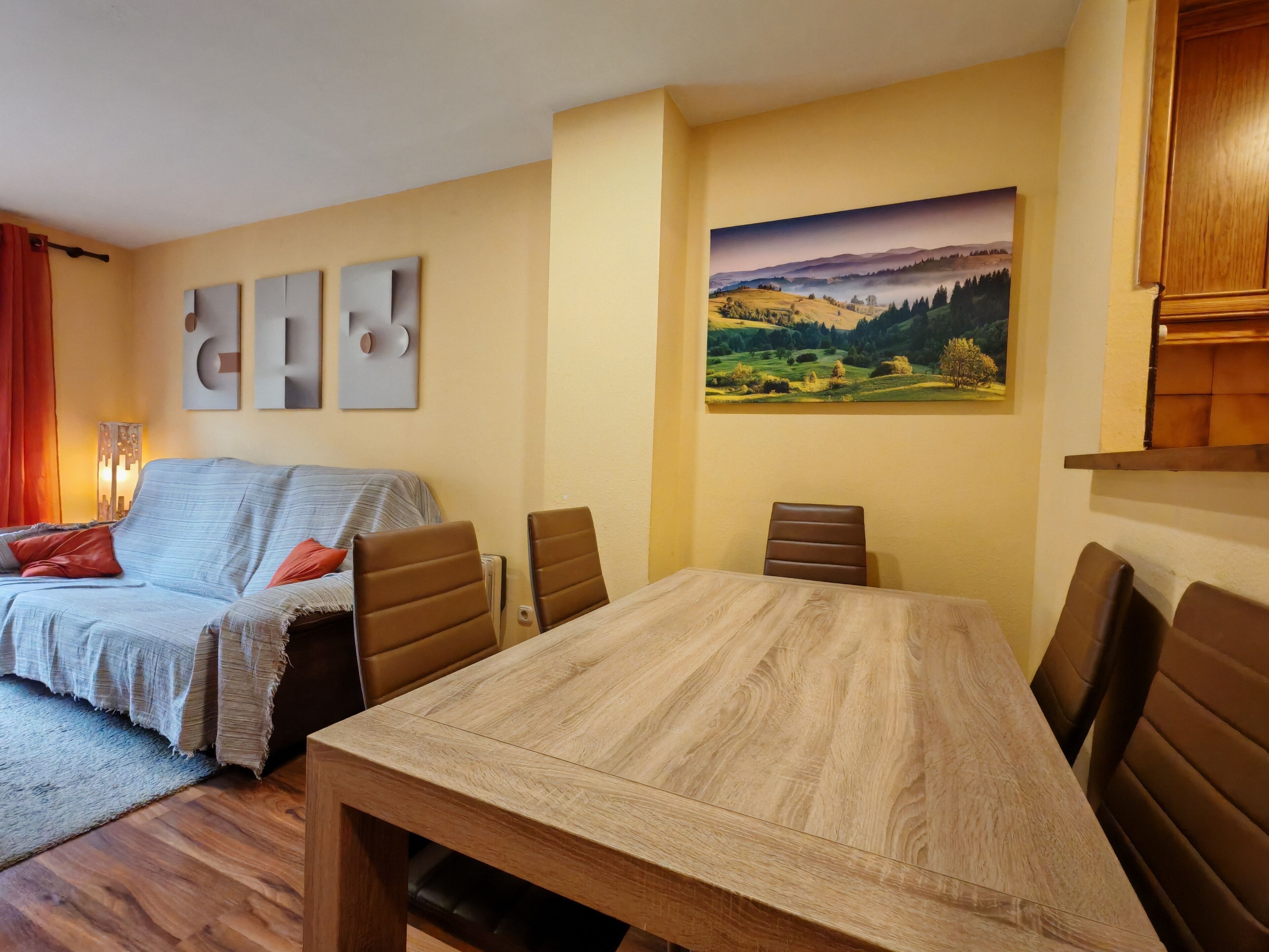 A cozy living room with a sofa, a dining table, and chairs. There is a painting on the wall and a lamp on the side.
