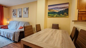 Una sala de estar acogedora con un sofá, una mesa de comedor y sillas. Hay un cuadro en la pared y una lámpara en la mesita.