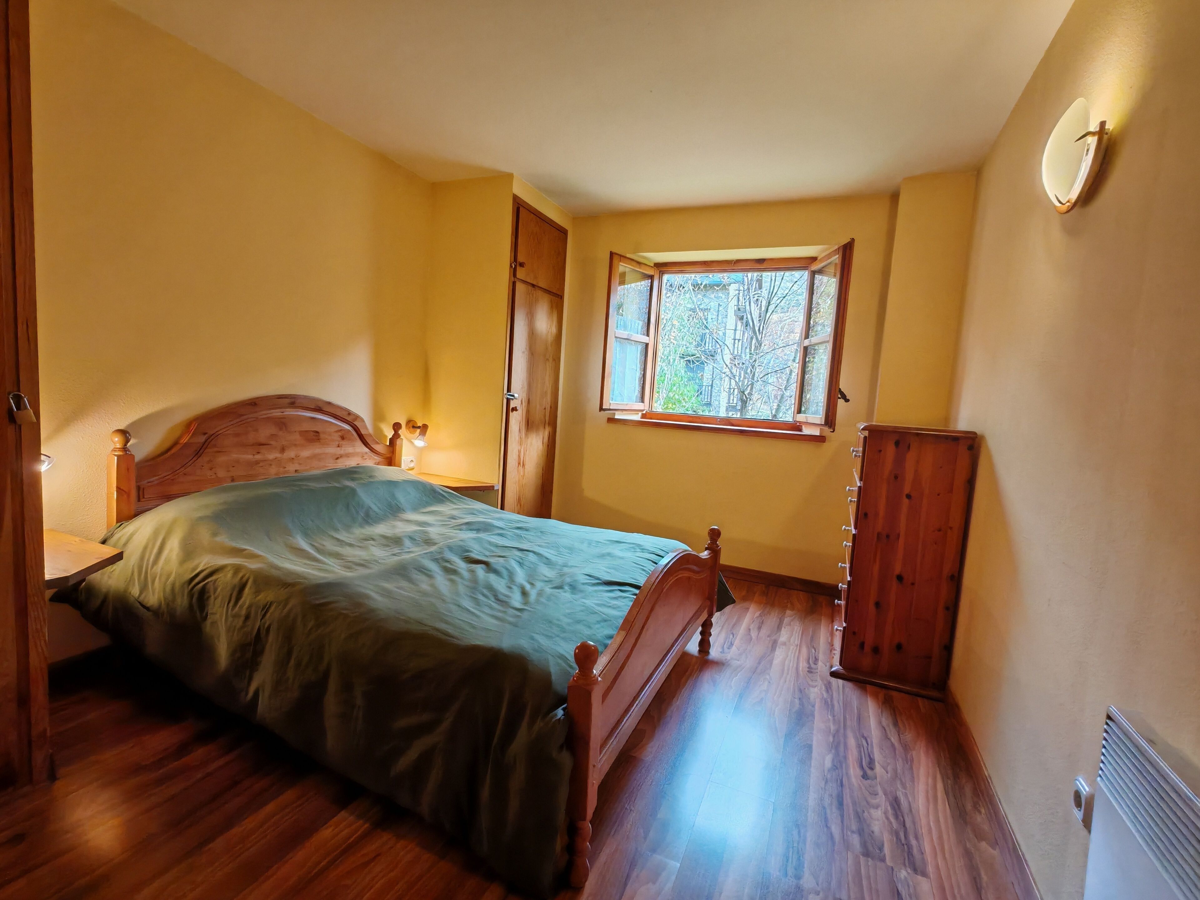 A bedroom with a wooden bed, a nightstand, a dresser, a window with a view of trees, and a radiator.