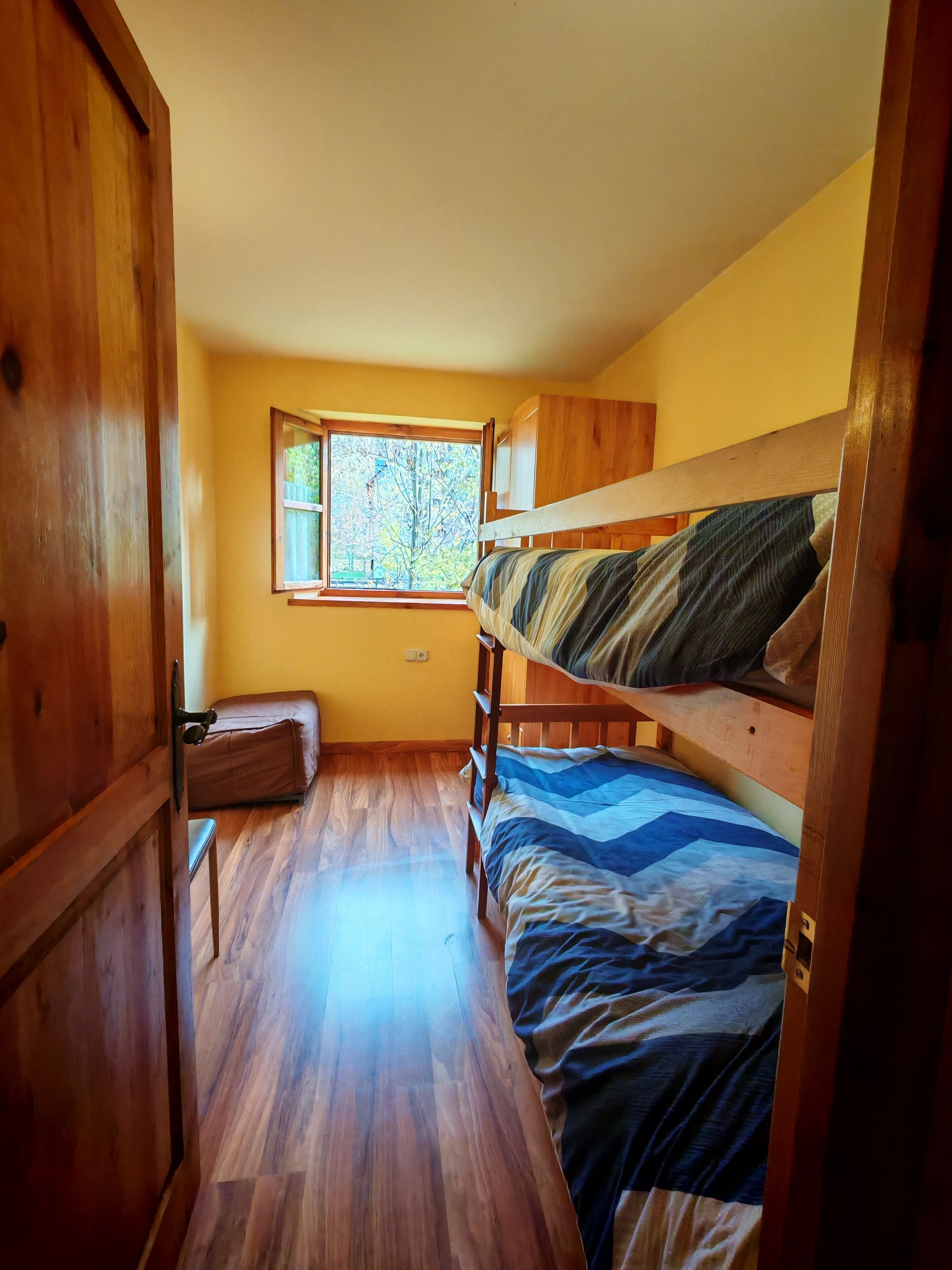 A room with wooden bunk beds, a window with a view of trees, and a door.