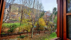 Vista a través de una ventana que muestra un cuarto con pared de piedra, puerta de madera y jardín con árboles y un edificio distante.