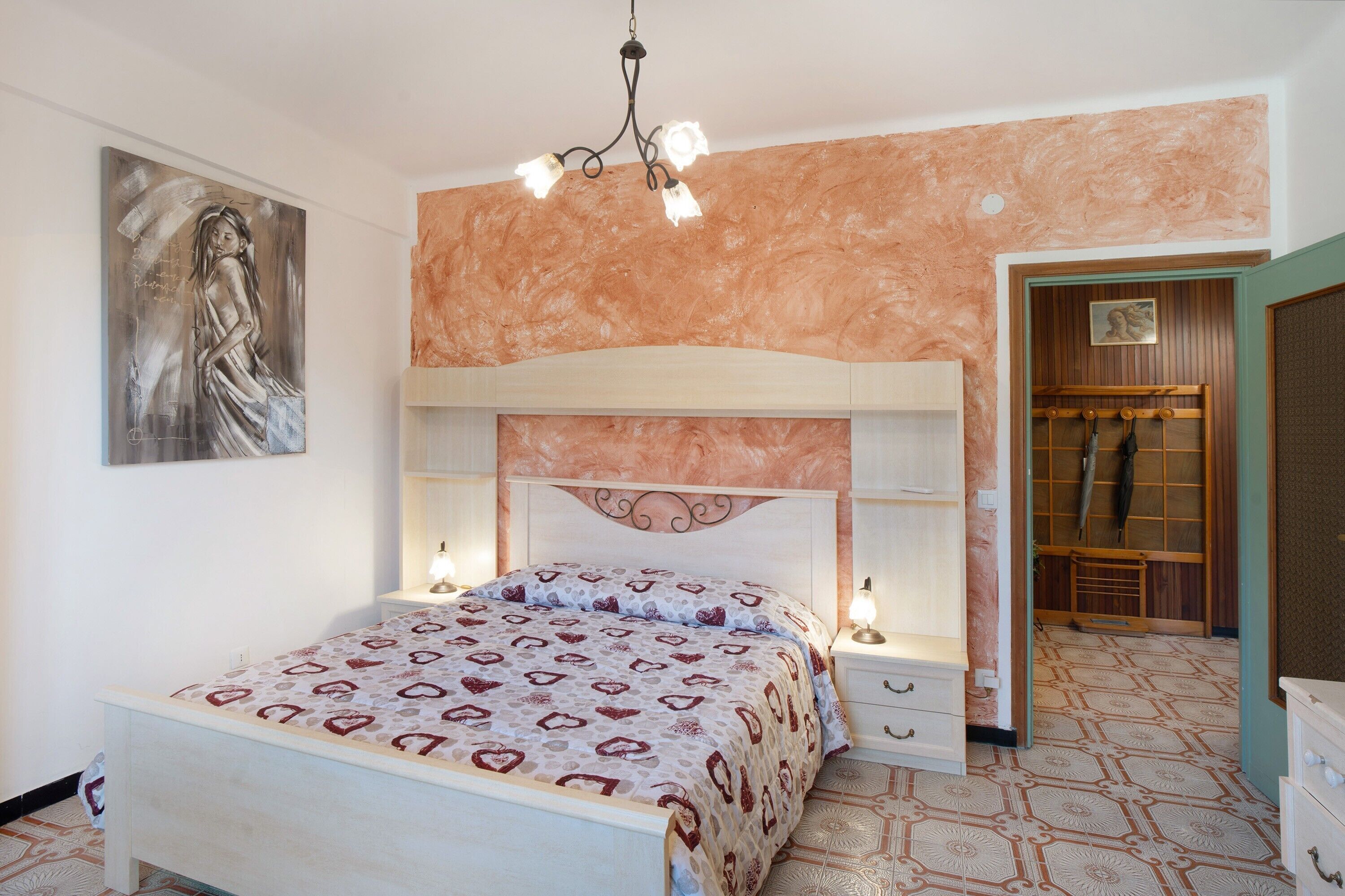 Una camera da letto con un letto, comodini, una lampada da comodino, un lampadario, un quadro appeso al muro e una porta in legno.