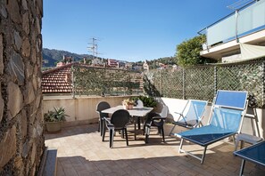 A patio with a table and chairs, a blue sun lounger, and a view of buildings and hills.