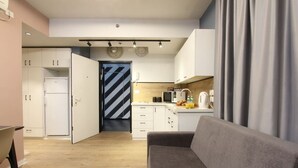 Eine moderne Küche mit weißen Schränken, einer Mikrowelle und einer Kaffeemaschine. Im Wohnbereich befindet sich ein graues Sofa und eine Tür mit Streifenmuster.