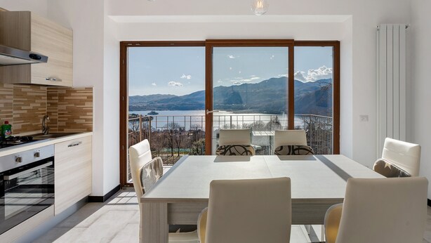 Une cuisine moderne avec une grande table à manger et des chaises, une vue sur les montagnes et un lac, ainsi qu’un balcon doté d’une balustrade.