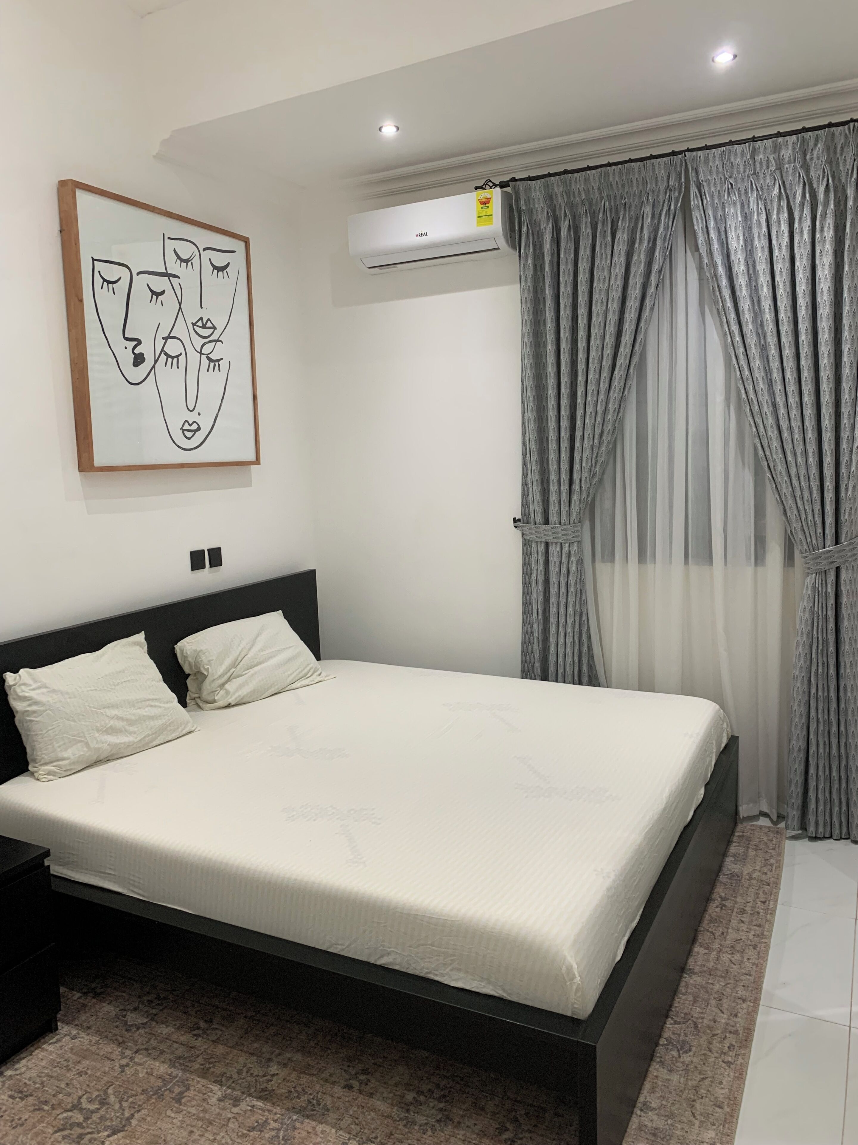 A bedroom with a bed, a headboard, a nightstand, a wall-mounted air conditioner, and a window with curtains.